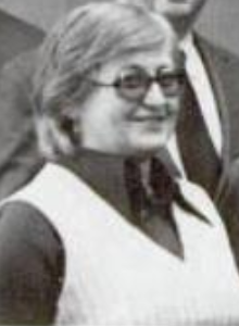 A smiling white woman with short grey hair, wearing tinted glasses, a dark blouse with a wide pointed collar, and a white sweatervest