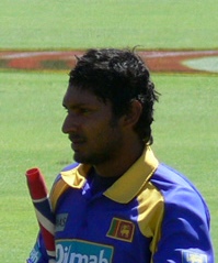 Portrait picture of a dark skinned man wearing blue and yellow Sri Lankan cricket kit, with a partially visible cricket bat under his arm.