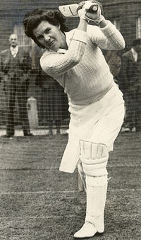 A full length black and white photograph of a female cricketer in pads playing a shot in the nets