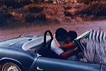A man and woman kiss in a convertible sports car, with the clapperboard accidentally at the edge of the frame.
