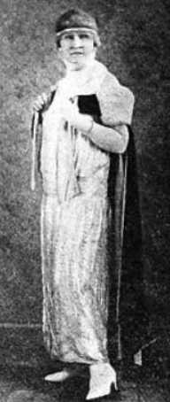 Full-length photo of a white woman, standing, wearing a long 1920s gown and headband, holding a book