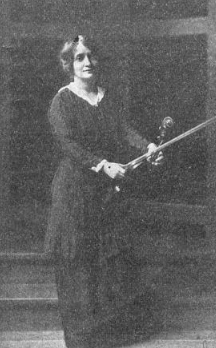 A white woman, standing, wearing a long dark dress, holding a violin and bow.