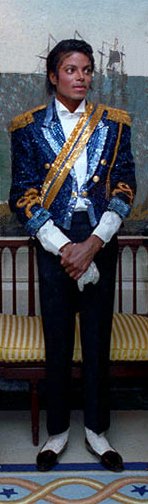 An African American man in his mid-twenties wears a sequined military jacket and dark aviator sunglasses. He walks and waves his right hand, which is adorned with a white glove. His left hand is bare.