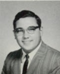 A black and white photograph from the shoulders up of Micheal D'Andrea, a pale skinned man with dark short hair and glasses, wearing a suit and tie