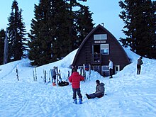 A dark brown building surrounded by trees in the background and snow, skis and people in the foreground.