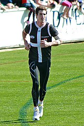 A male athlete with dark hair wearing a sleeved jersey and compression pants runs on the boundary of the grass surface of the playing arena.