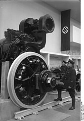 A traction motor from a German E18 class at an industrial exhibition. There is a Nazi swastika flag hanging behind and a man in military uniform is striking a heroic pose as if admiring the motor.