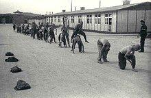 A line of half-naked prisoners performing "leap frog", under supervision of one of the Kapos. In the background the main gate to Mauthausen as well as two wooden barracks are visible.