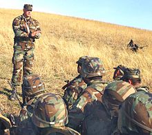 A soldier standing in a field, speaking to other soldiers sitting on the ground