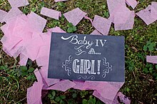 A black sign with white handwritten letters reads "Baby IV is a...girl!" The sign rests on grass and is surrounded by pieces of pink crepe paper confetti.