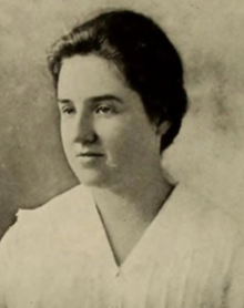 A young white woman with dark hair dressed back to the nape, wearing a white blouse (might be an academic gown)