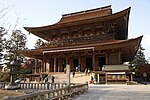 Large wooden building with a hip-and-gable roof and an enclosing pent roof. There is a railed open veranda above the pent roof.