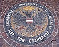 A mosaic laid into stone. It is not visible, but it lays in front of the University Church. It displays the old crest of Albert Ludwig, the leader who instituted the University. It displays a two-headed eagle in dark stone with an austrain crest in the middle, surrounded by brown stones, surrounded by a ring of dark stone and white stone text.