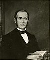 White male with dark hair and long sideburns in a dark suit sitting at a desk with an open book