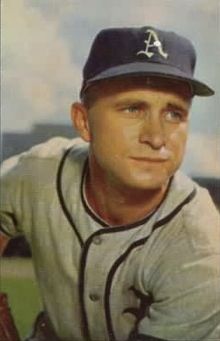 A man wearing a white baseball uniform and a navy-blue cap with a blackletter "A" on the face in white