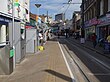 A paved road with one tram track in the middle and a tram shelter on the left with many street-level shops on the right hand side.