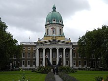 A building with white pillars and a white and green-domed ceiling, with the words "Imperial War Museum" beneath the dome and behind a flagpole. Cannons sit in on the lawn in front of the building, and a group of people stand on sidewalks that cut across the grassy lawn.