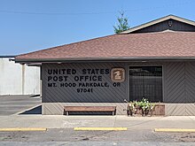 Front-on photo of a brown post office building with a sloped roof, standing alone in a bland parking lot, with large letters on the wall reading "United States Post Office Mt. Hood Parkdale, Oregon 97041" and a golden "United States Postal Service"/"US Mail" logo.