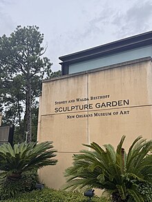 Entrance to the Sydney and Walda Besthoff Sculpture Garden at the New Orleans Museum of Art, with the garden's name displayed on a beige wall, surrounded by green plants and trees.