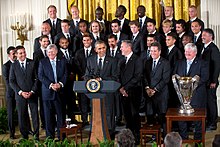 A group of soccer players for Sporting Kansas City dressed in suits behind President Barack Obama, who stands at a podium. The MLS Cup trophy stands to the President's left on a table.