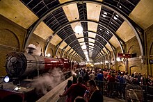 On the left is a red steam locomotive and three carriages, with steam drifting out above. On the right passengers queue behind barriers and signs that say "Hogwarts Express" and "Platform 9¾". Over both of these is a large cylindrical glass and steel roof.