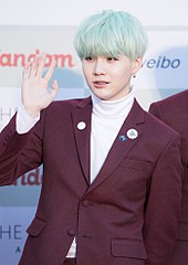 Suga, with blue hair and in a pink suit, holds his right hand up and looks left