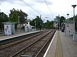 A set of two tram tracks between two platforms with shelters. There is a level pedestrian crossing just before the horizon.