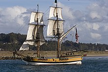 A white and gold antique wooden sailing ship sits in a body of water, with land behind it.