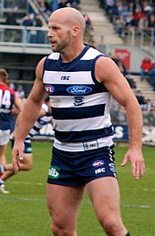 A male bald-headed athlete wearing a sleeved jersey and shorts standing on the grass surface of the playing arena.