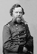 Black and white photo shows a bearded man with an unusually long moustache. He wears a dark military uniform with the shoulder tabs of a major general.