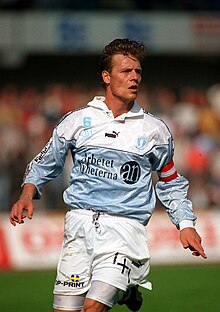 A broad-shouldered, Nordic-looking association football player, pictured mid-match. Looking to his left (the viewer's right), he sports a sky blue, white and black shirt and white shorts, and a red and white striped band around his left arm.