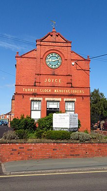 Colour photograph showing the former factory of J. B. Joyce & Co. Constructed of red brick and shown from the front. A large clock is positioned above a window with the wording "Joyce, Turret Clock Manufacturers", in capital letters, affixed to the frontage.