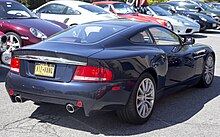 A rear-three quarters view of a very dark blue Vanquish outdoor, with some Porsches parked in the background.