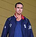 An English cricketer with closely cut dark brown hair, in a dark blue jacket. He is wearing a blue T-shirt and has sun-cream on his cheeks.