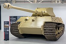 A frontal view of a large, pale-yellow tank in a white museum gallery.. Its curved-faced turret is pointing forwards, the long gun overhangs the front by several meters.