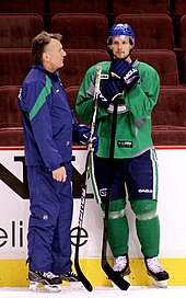 A Caucasian ice hockey player skating with gripping a hockey stick with both hands. He wears a green jersey with white and blue trim and a blue, visored helmet.