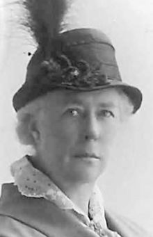 An older white woman with white hair, wearing a cap with a plume, and a blouse with a lace collar