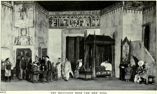 A tall stage-set depicting a large room with medieval pictures, patterns and motifs on the upper parts of the walls. On the left, there is an alcove and two sets of doors. At the back is a tall screen and an elaborate curtained four-poster bed containing a man in sleeping garments and a nightcap. To the right is a tall wooden desk with a religious picture in a gothic frame, sideways-on to a short staircase leading up to a balcony door. Seven men (two of them tradesmen), three women and a child, all in medieval garments, are standing or sitting around the room listening to an important-looking man who is reading a document out loud.