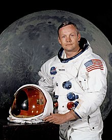 A light-skinned man in his late 30s, with blue eyes and brown hair parted to the right. He wears a white space suit, and holds the helmet. The space suit has five hose connectors on the front. There is a large U.S. flag on the left shoulder. The helmet's transparent faceplate is tinted gold.