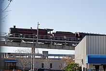 The lower half of the photograph shows industrial office buildings, with a small label "T-4" in one corner. Above this runs a steel construction carrying an elevated rail system, with an emergency exit walkway. Mounted on the track is a red steam locomotive with two small wheels and three big wheels raised slightly above the track. Above the middle of the three big wheels is a curved sign that says "Hogwarts Castle". Under the train are sets of much smaller wheels resting directly on the track.