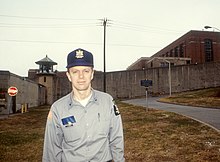 Ted Conover in his late 30s, standing outside of the prison perimeter wall wearing his correctional officer uniform hat and button-up shirt, with officer's notebook and pen in his shirt pockets