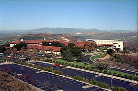 A complex of buildings is in the middle ground. Behind it, a view of a valley.