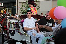 A young man in a white tee-shirt with ripped jeans and a man in a black tee-shirt that reads "Annie's" ride in a white horse-drawn carriage.