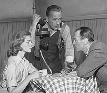 Lauren Bacall in black and white sitting at a table with Humphrey Bogart and Henry Fonda while performing a scene on an episode of The Petrified Forest in 1956