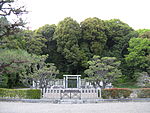 Concrete torii gate behind two concrete fences and in front of a group of trees.