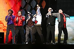 Six men wearing multicolored clothes are seen performing onstage.