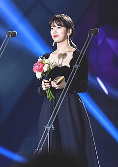 Bae Suzy wearing a long black dress on stage, smiling while holding a bouquet and an award