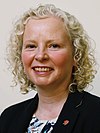 Official Portrait of Claire Baker MSP. She is smiling, with curly blonde hair and a blazer with a rose pin. There is a grey-ish background.