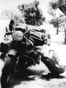 Black and white photo of woman crouched down next to a motorbike