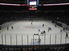 An ice hockey rink on which two teams are playing seen from some distance up and behind the goal. High transparent shielding mounted on the wall surrounds the rink.